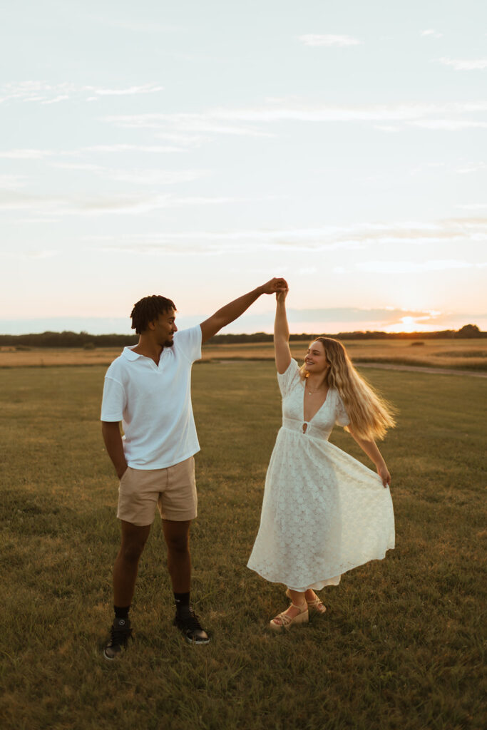 A couple twirls in a field as the sunsets during their summer engagement photos