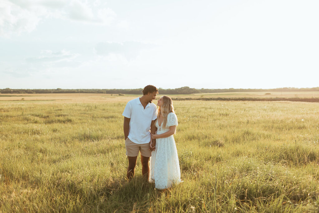 A couple stands in a field while they gaze at each other during their engagement session at the barn at Grace Hill